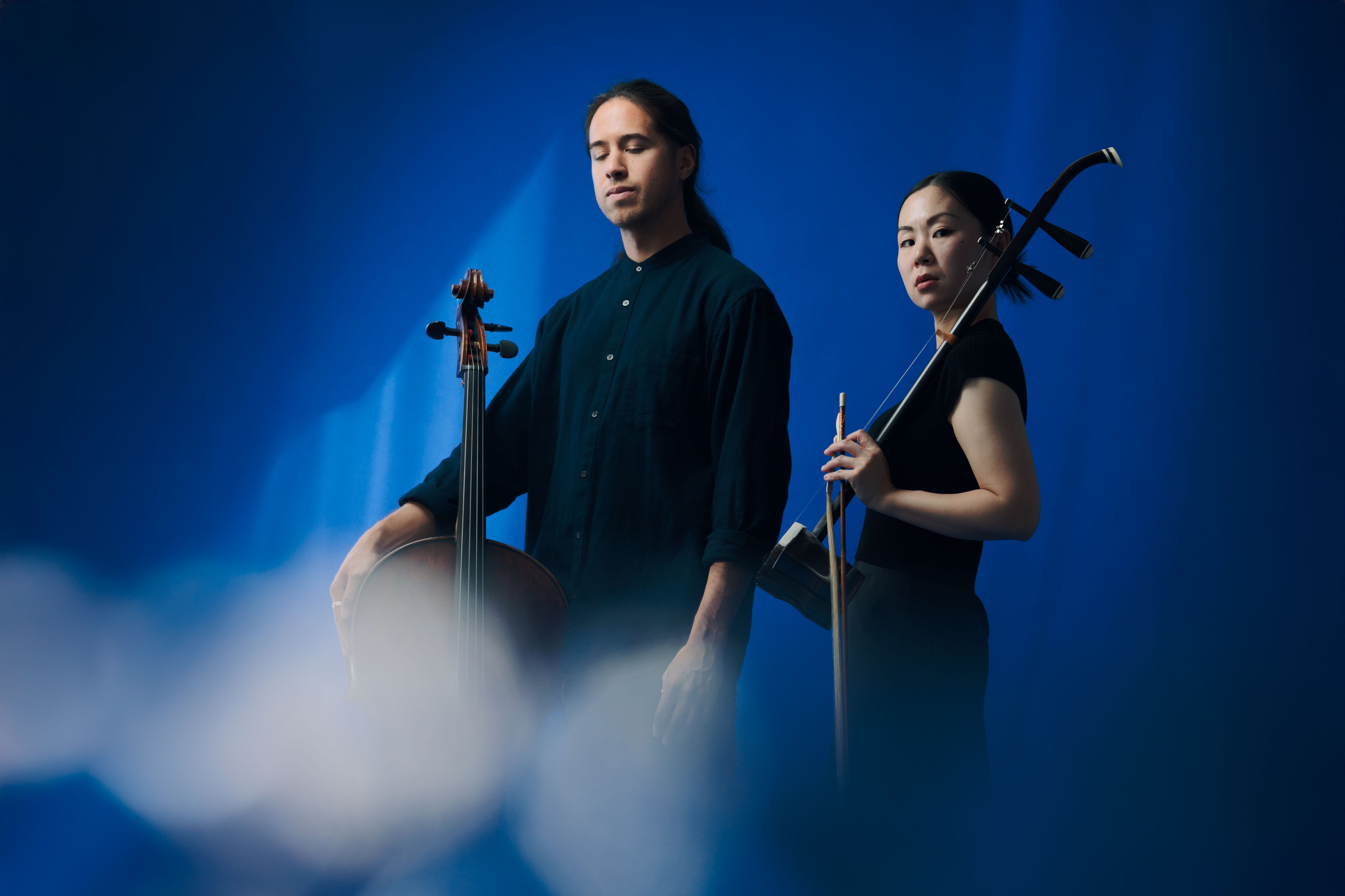 Two musicians, a man with a cello and a woman with an erhu, pose thoughtfully against a vibrant blue backdrop filtered through soft, white light.