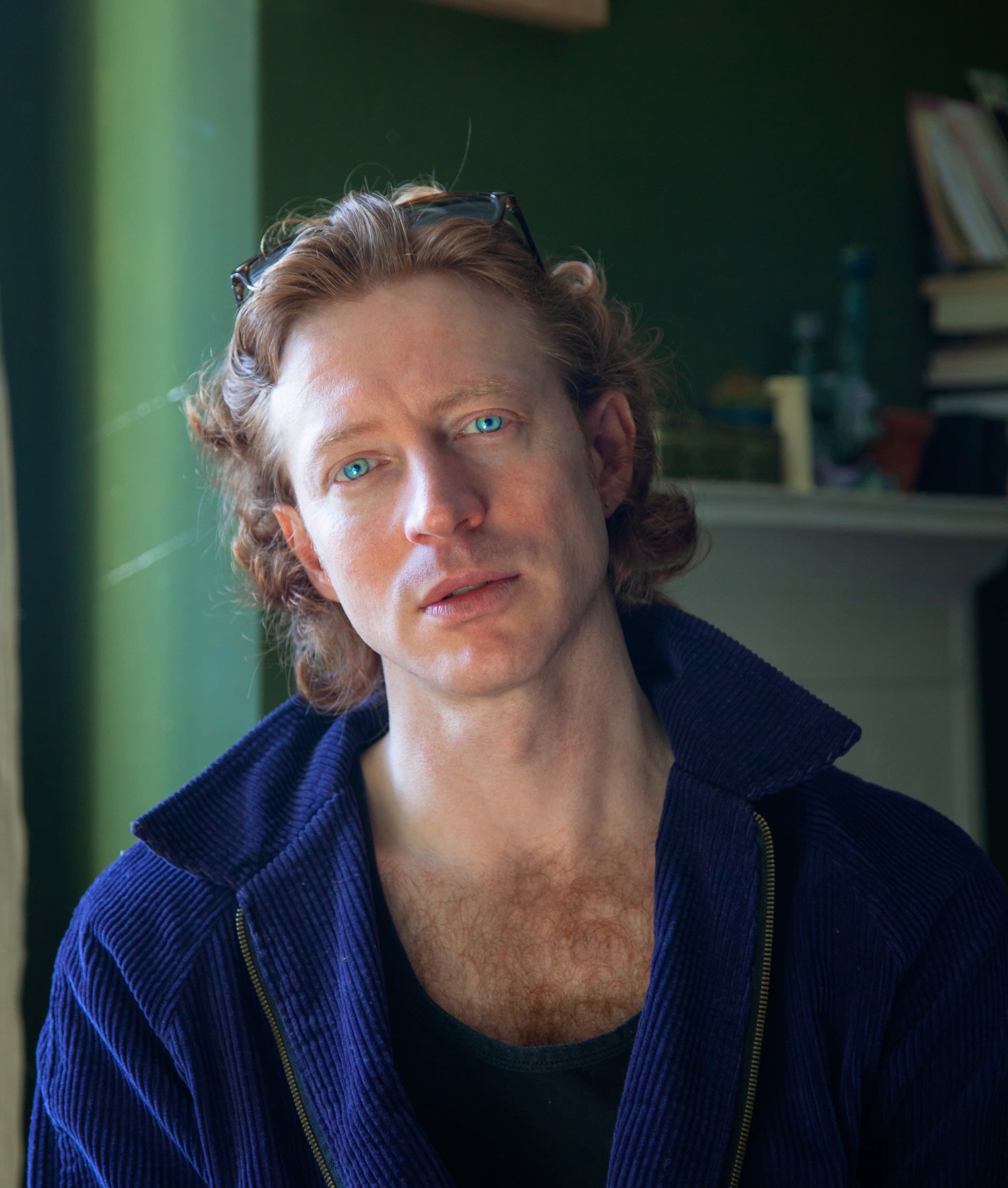A close-up, dramatic portrait of Cameron Graham with wavy red hair and piercing blue eyes looking directly at the camera while wearing a blue corduroy zip-up jacket.