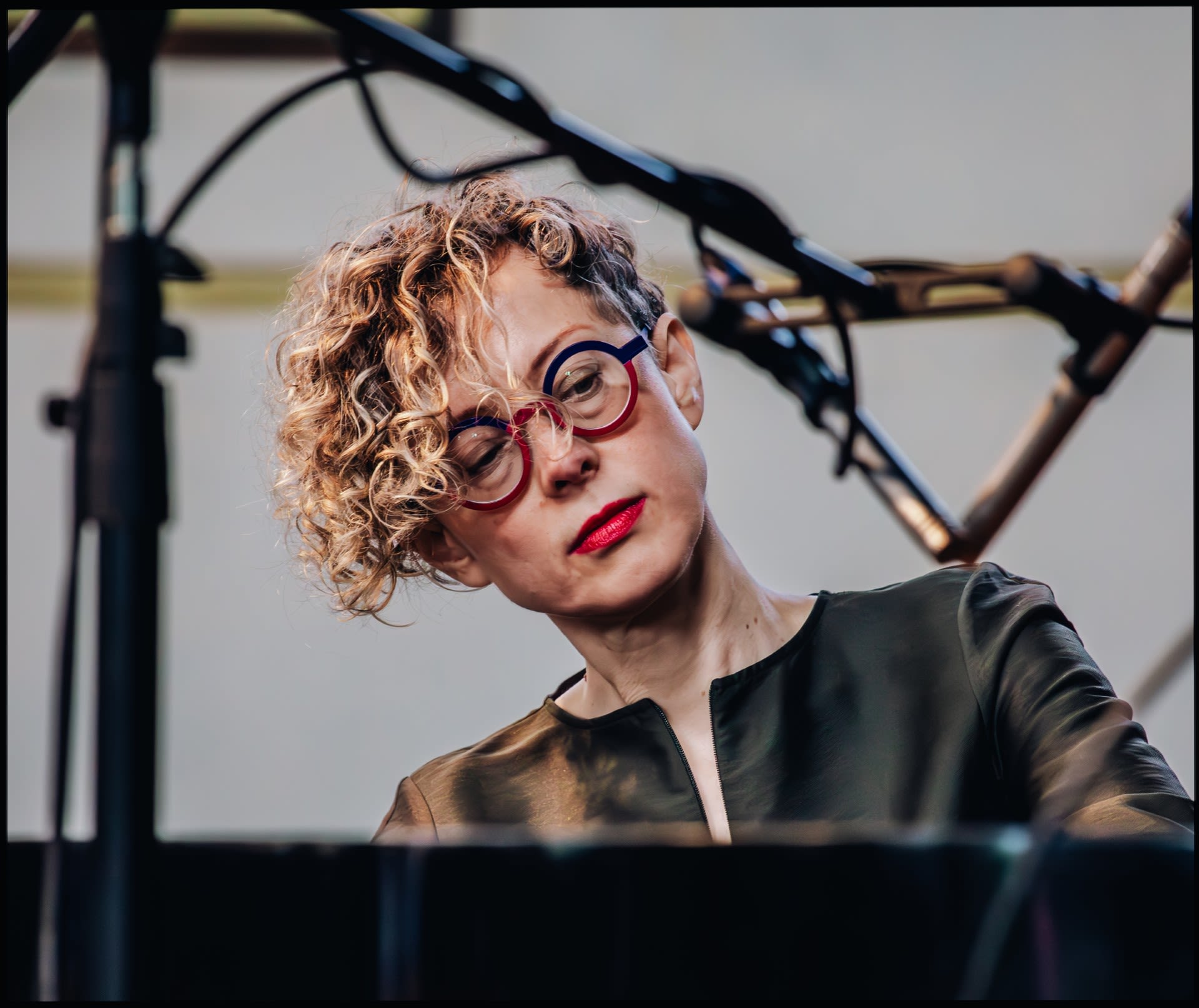 Xenia Pestova Bennett  with curly blonde hair and distinctive red-and-blue circular glasses looks down while playing a piano.