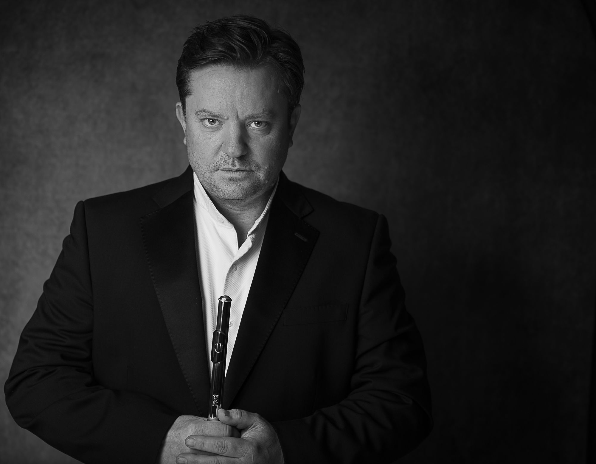Gareth is holding a flute in his hand. He is wearing a black and white suit with a white shirt underneath, and his hair is neatly combed. His expression is serious and focused, as if he is deep in thought.