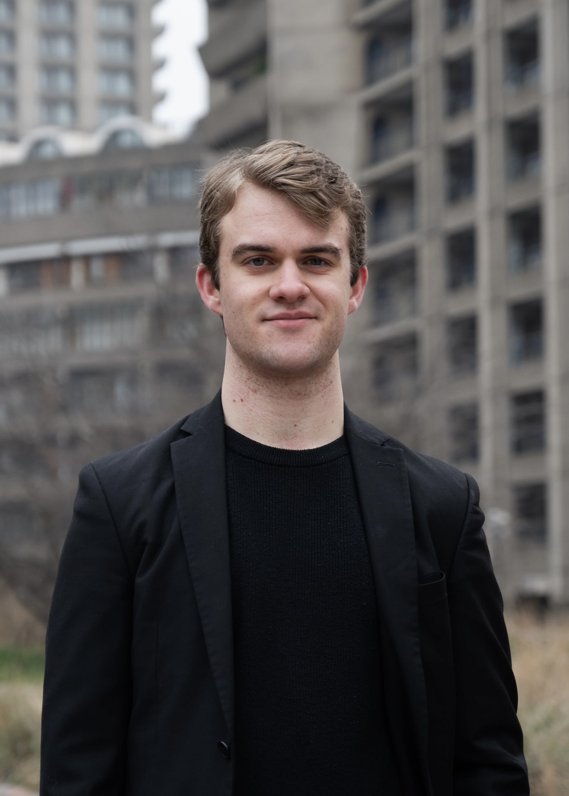 Owen Lee standing in front of a tall building, surrounded by grass and a clear blue sky. He is wearing a black suit.