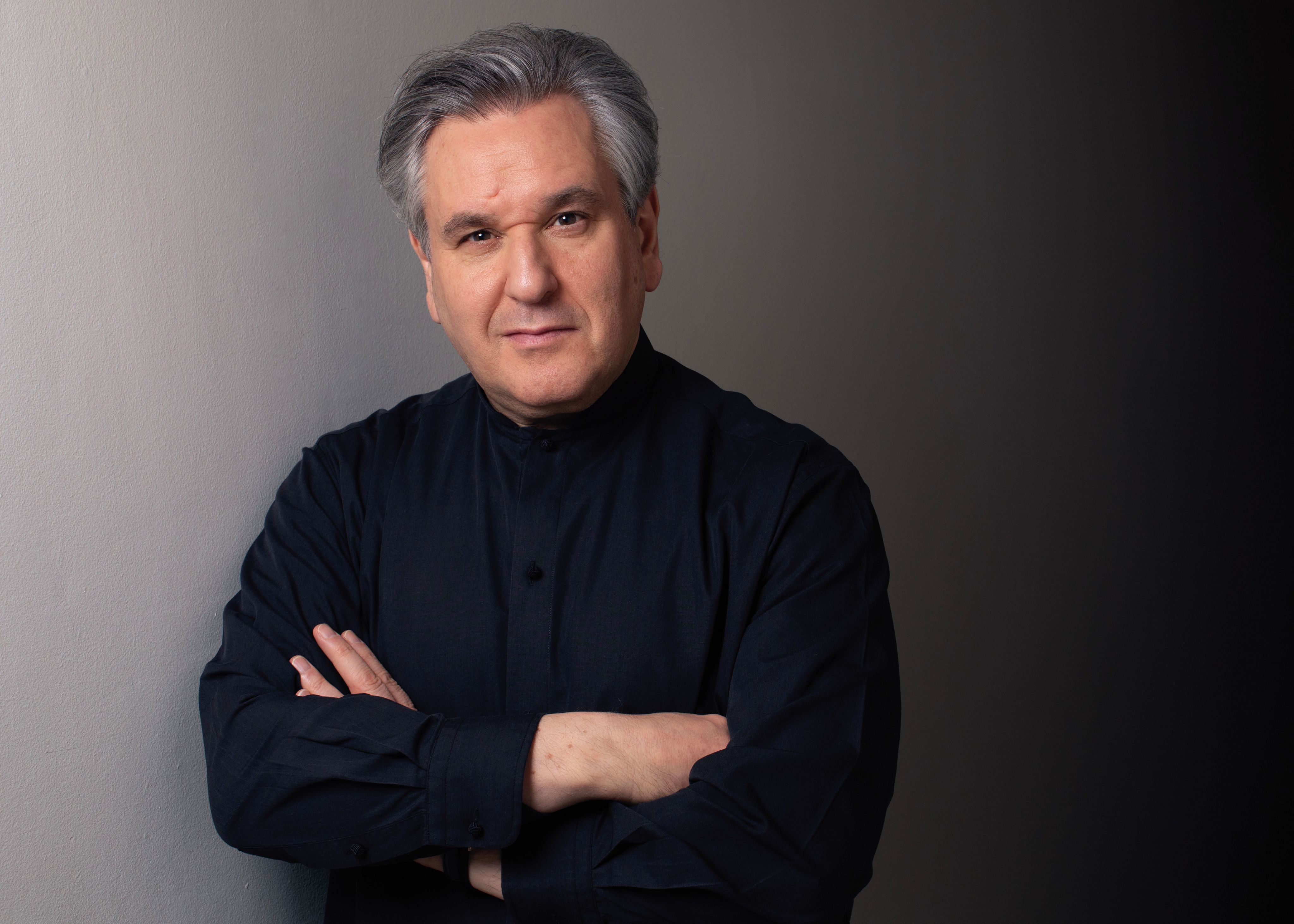 A portrait of conductor Sir Antonio Pappano, featuring him with grey hair, dressed in a dark blue shirt, and posing with his arms crossed against a grey background.