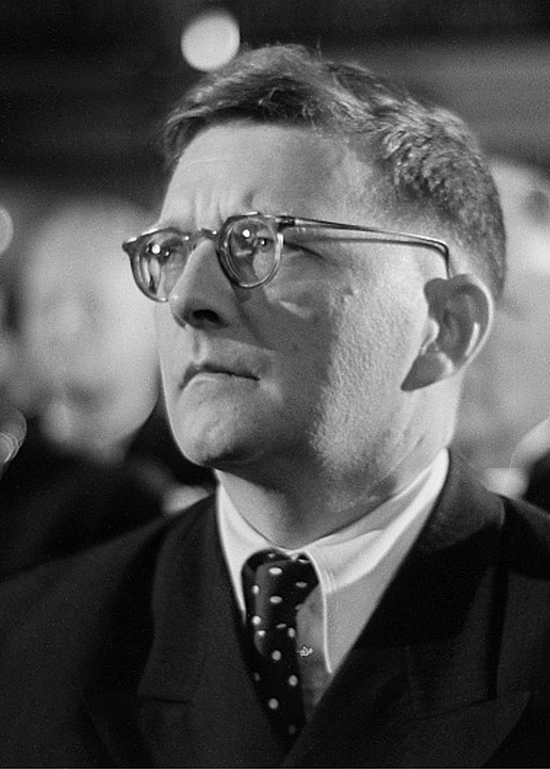 A black-and-white portrait of Russian composer Dmitri Shostakovich, wearing round glasses, a suit, and a polka-dot tie.